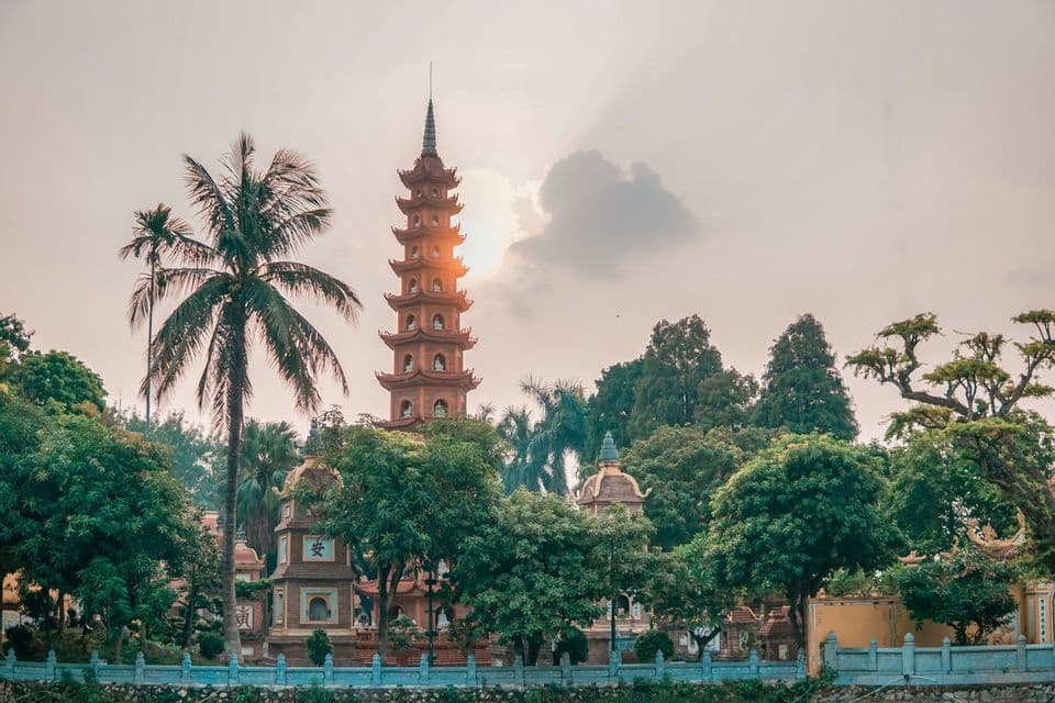 Una pagoda de varios niveles se alza entre frondosos árboles verdes y una palmera mientras el sol se pone detrás de ella.