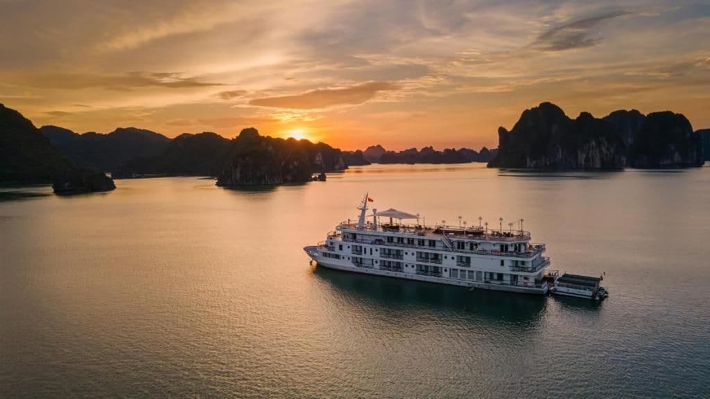 Un paquebot blanc flotte sur une eau calme, entouré d'îles rocheuses au coucher du soleil.