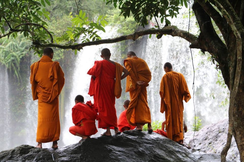 Un groupe de moines bouddhistes, vêtus de robes orange et rouges, se tient sur un rocher mouillé, observant une grande cascade dans une forêt luxuriante.