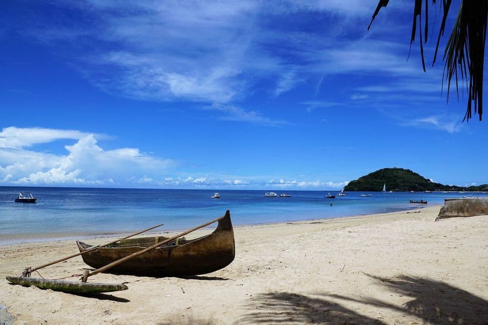Una canoa a bilanciere in legno si trova su una spiaggia di sabbia bianca, con un mare calmo e turchese e un'isola verde sullo sfondo.