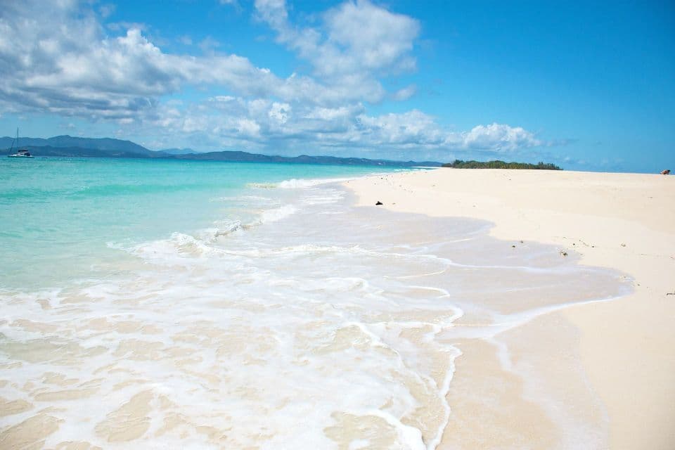 Una vasta spiaggia di sabbia bianca si estende lungo un litorale turchese sotto un cielo azzurro con nuvole sparse e montagne sullo sfondo.
