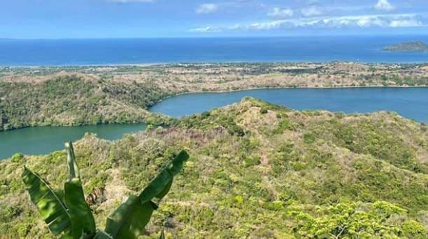Vista dall'alto su un rigoglioso e verde cratere vulcanico con un lago e l'oceano blu visibile sullo sfondo.