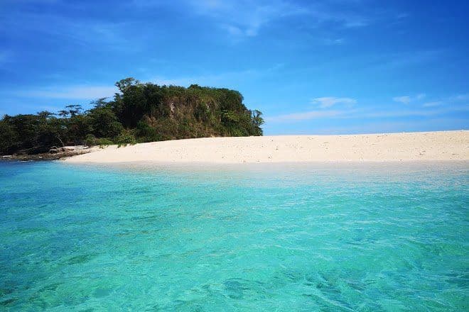 Una spiaggia di sabbia bianca deserta su una piccola isola tropicale con acqua turchese in primo piano e un cielo azzurro sopra.
