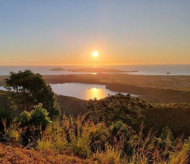 Una vista panoramica da una collina al tramonto, che domina un lago calmo, colline verdi e l'oceano con isole all'orizzonte.