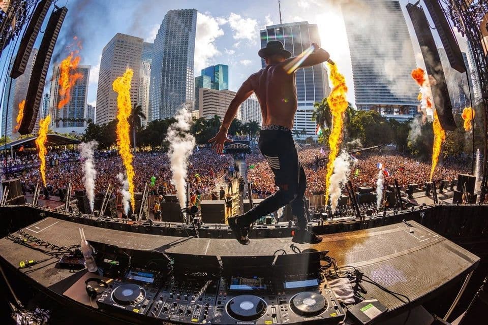 Un artista a torso nudo con un cappello salta su un palco con fuochi d'artificio di fronte a una folla immensa a un festival musicale all'aperto.