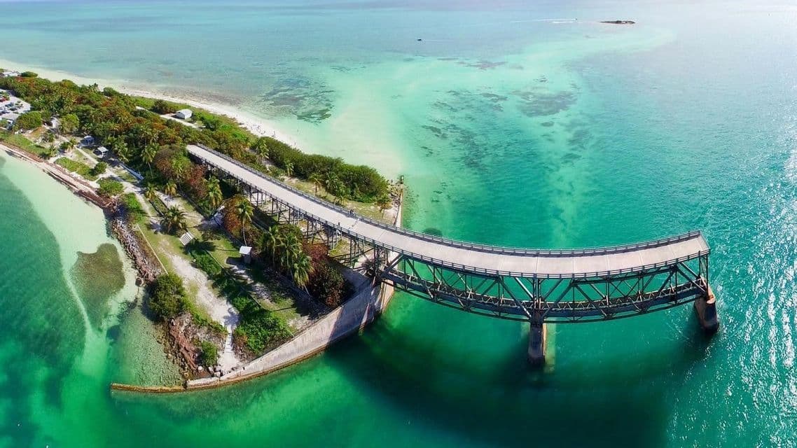 Una vista aerea di un lungo ponte curvo che si estende da una penisola verde su acque oceaniche limpide e turchesi.
