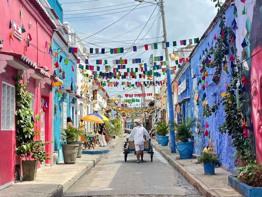 Un homme pousse un chariot dans une ruelle étroite bordée de bâtiments colorés et de drapeaux festifs suspendus.