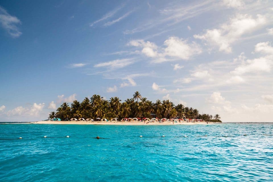 Une île tropicale avec des palmiers et des tentes colorées sur une plage de sable, vue de la mer turquoise sous un ciel partiellement nuageux.