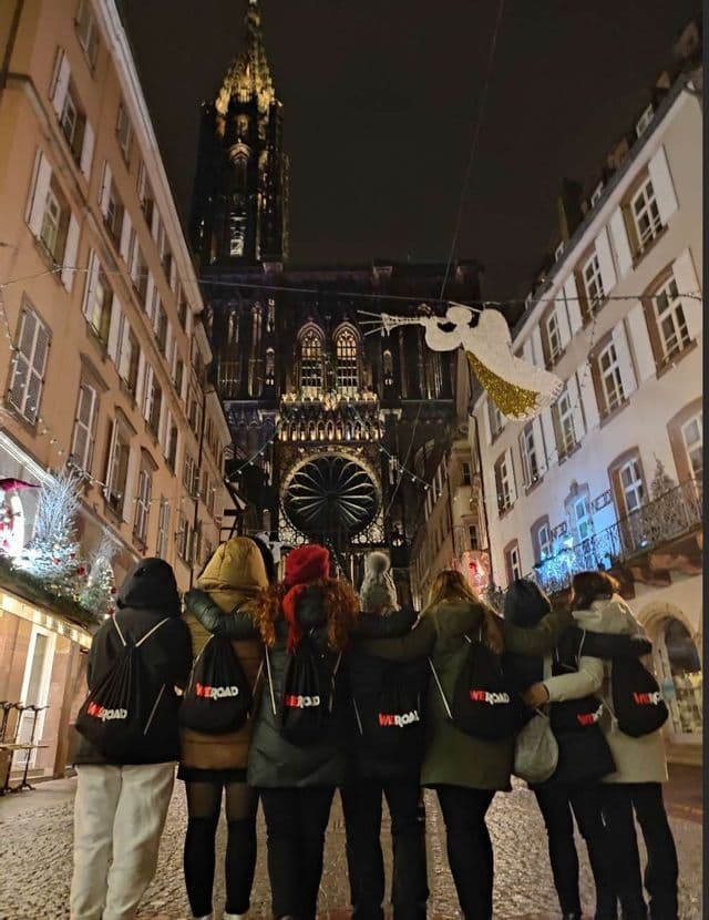 Un gruppo WeRoad si trova insieme in una strada di notte, di fronte a una cattedrale gotica illuminata con decorazioni natalizie.