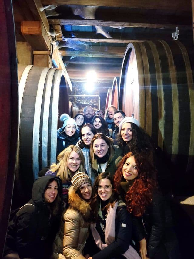 Un viaggio di gruppo WeRoad con persone che sorridono per una foto in una stretta cantina tra grandi botti di vino in legno.