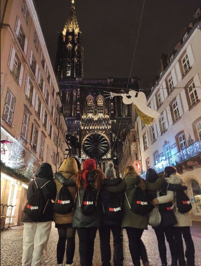 A WeRoad group trip seen from behind, standing arm-in-arm on a decorated cobblestone street at night, looking at a large illuminated cathedral.