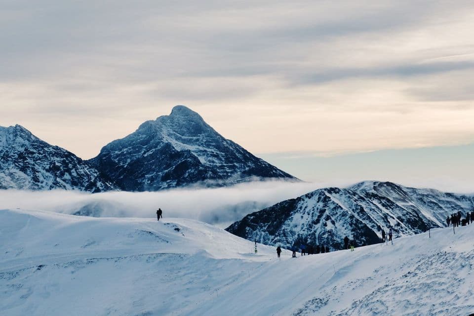 Eine WeRoad-Gruppe steht auf einem verschneiten Hang und blickt auf schroffe Berggipfel, die über einer dichten Wolkendecke aufragen.