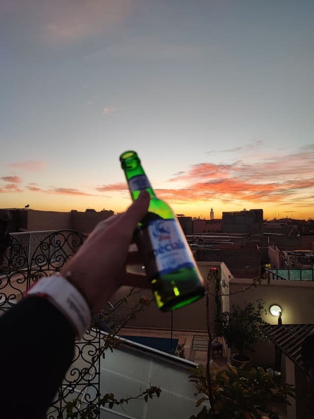 Una mano tiene una bottiglia di birra verde contro un cielo colorato al tramonto sopra i tetti di una città.