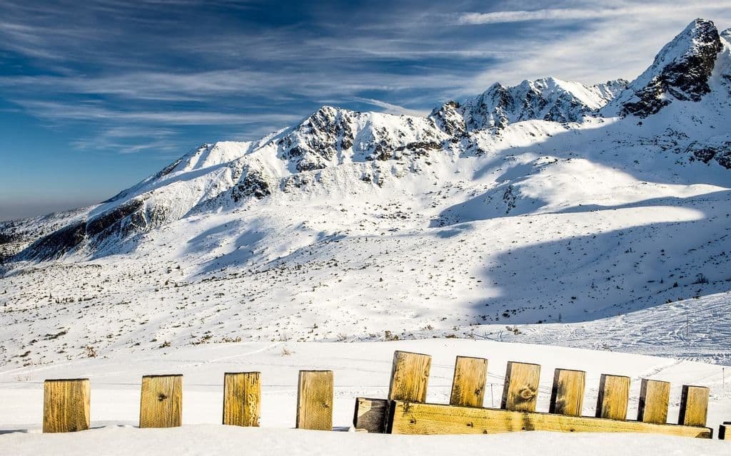 Ein zerbrochener Holzzaun ragt aus tiefem Schnee hervor, mit einem weiten, sonnenbeschienenen Gebirge im Hintergrund unter blauem Himmel.