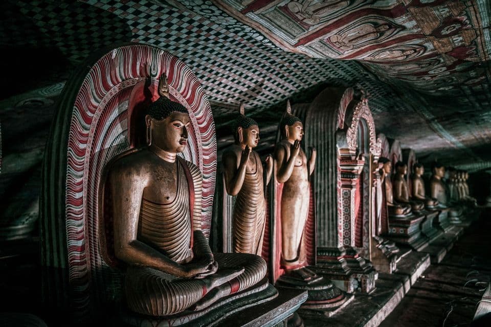 Una lunga fila di statue di Buddha scolpite all'interno di un tempio in grotta poco illuminato, con un soffitto riccamente decorato.