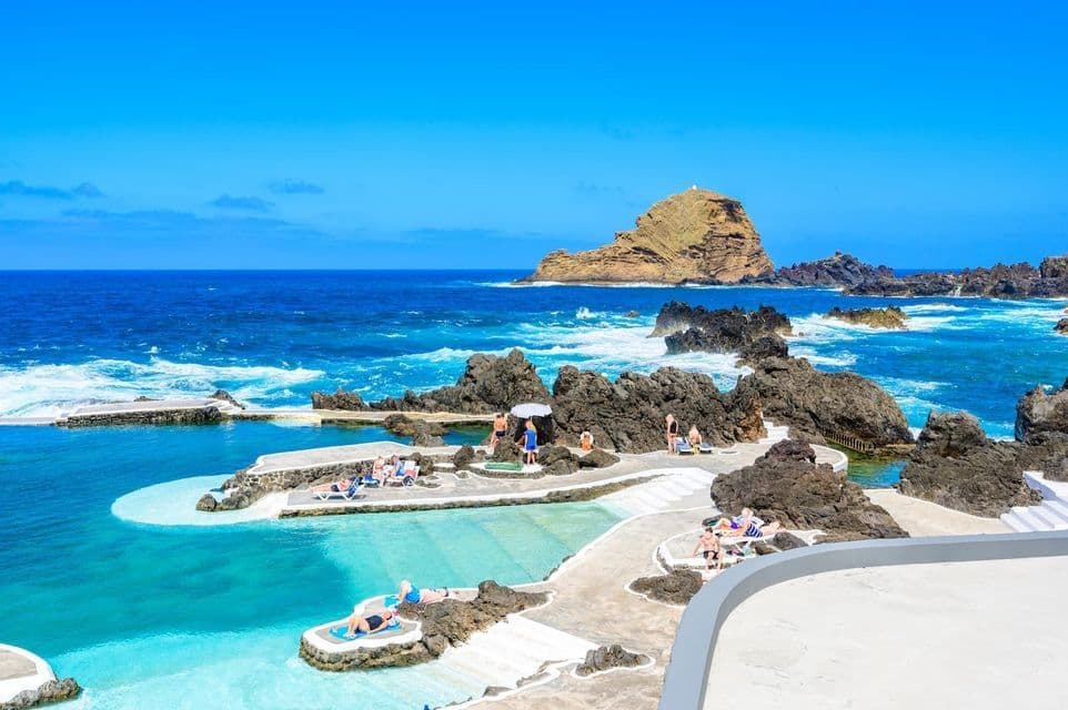 People sunbathing by natural lava rock swimming pools next to a deep blue ocean under a clear sky.