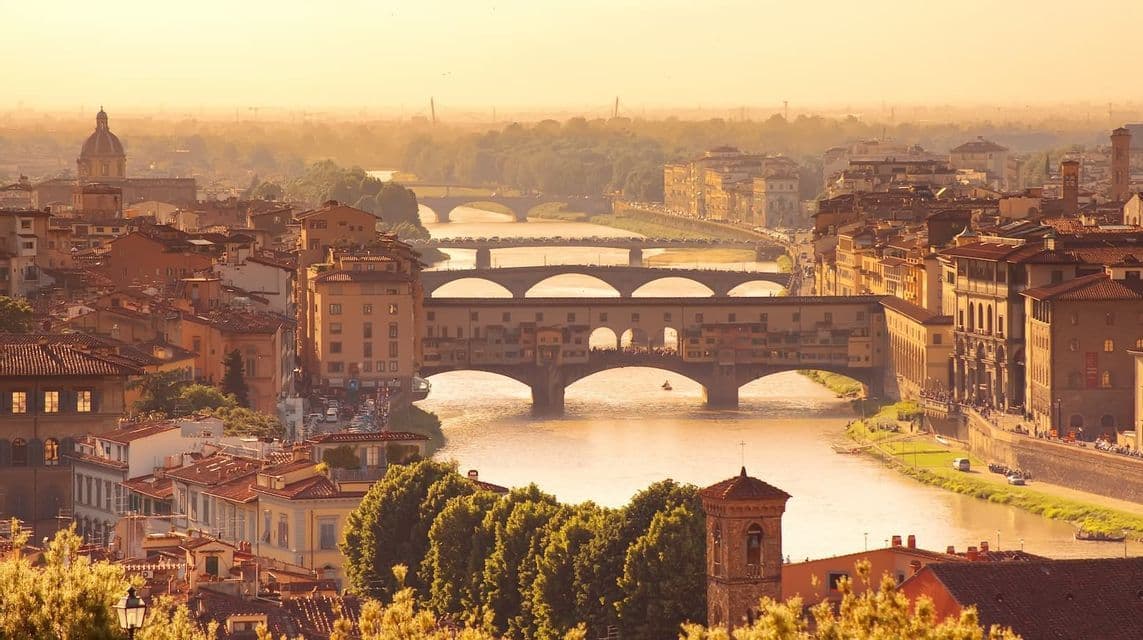 An aerial view of a historic city with terracotta roofs and a river crossed by several bridges during a golden sunset.