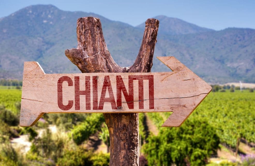 Un cartello in legno a forma di freccia indica a destra con la parola 'CHIANTI' in rosso, su uno sfondo di vigneti e montagne.