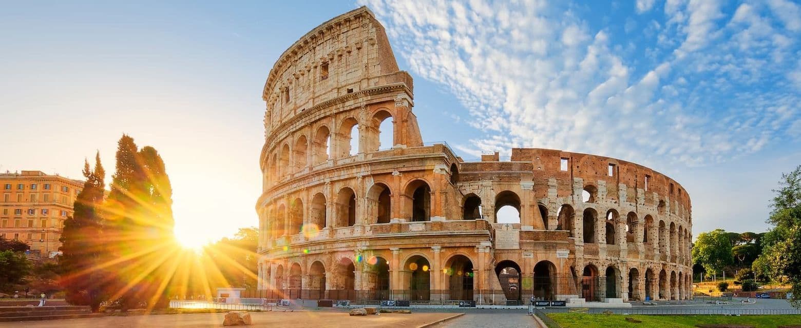 The ancient Colosseum stands illuminated by a bright sunburst against a blue sky with scattered clouds.