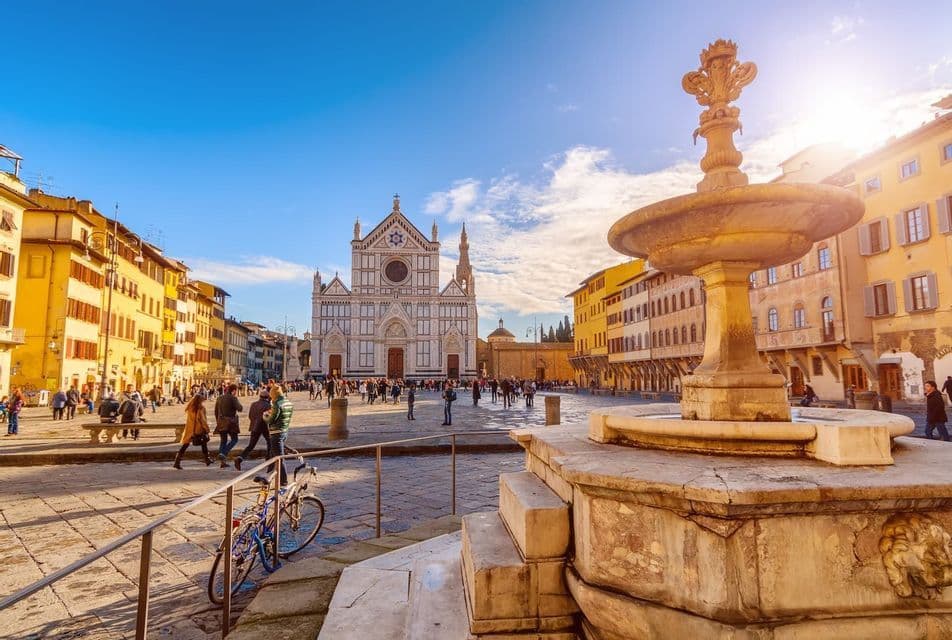 Una fontana in pietra in primo piano in una piazza europea soleggiata, con persone che camminano di fronte a una basilica bianca decorata.