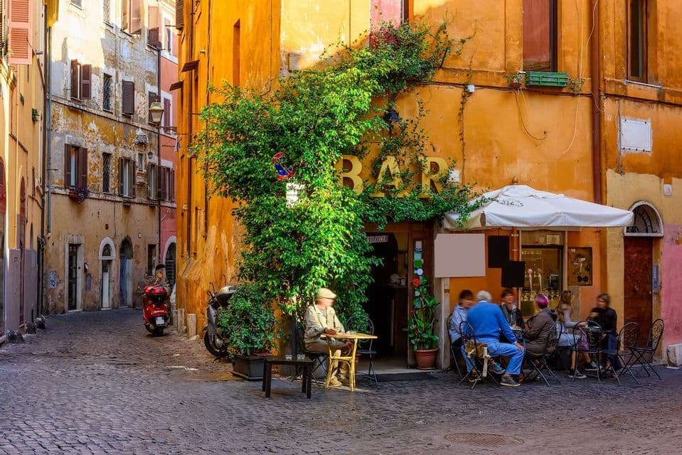 Une ruelle pavée ensoleillée avec des gens assis à un bar extérieur couvert de lierre verdoyant.