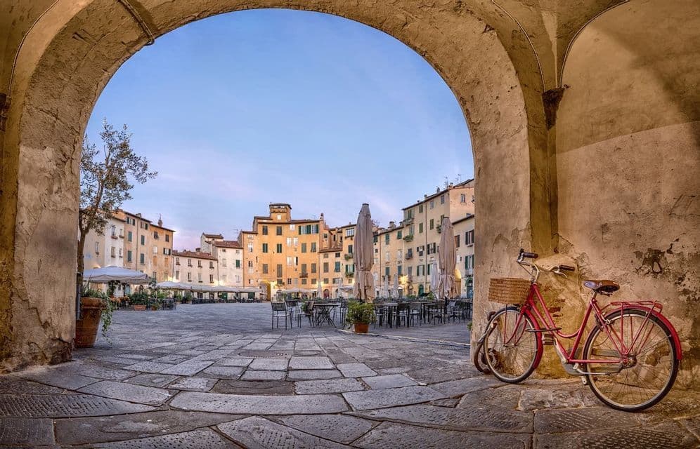 Una bicicleta roja con cesta está aparcada bajo un arco de piedra, con vistas a una plaza histórica adoquinada al anochecer.