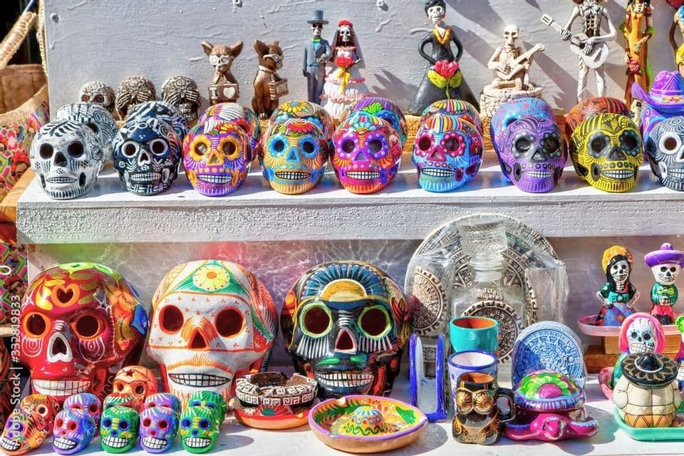 Teschi e statuette in ceramica colorati per il Giorno dei Morti, esposti su scaffali bianchi in un mercato.