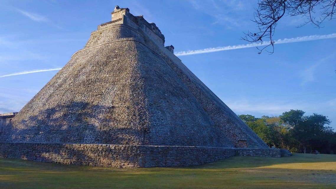 Una grande piramide di pietra stagionata con angoli arrotondati si erge su un prato verde sotto un cielo azzurro chiaro.