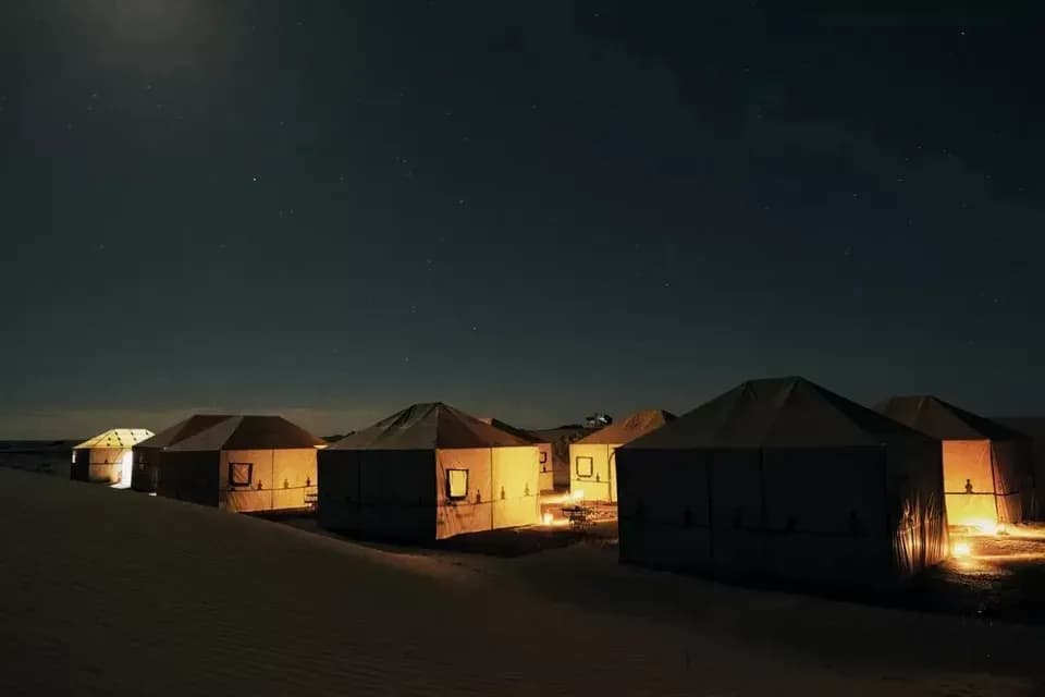 Una fila di tende di tela illuminate allestite su dune di sabbia sotto un cielo notturno scuro e stellato.