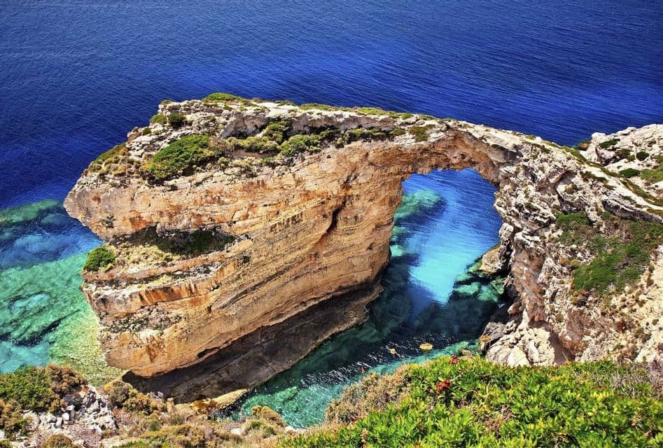 Ein natürlicher Steinbogen spannt sich von einer felsigen Klippe über klares türkisfarbenes Wasser, mit dem tiefblauen Meer im Hintergrund.
