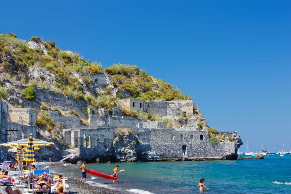 Una spiaggia di ciottoli assolata con persone che nuotano e si rilassano accanto a rovine in pietra incastonate in una verde collina.