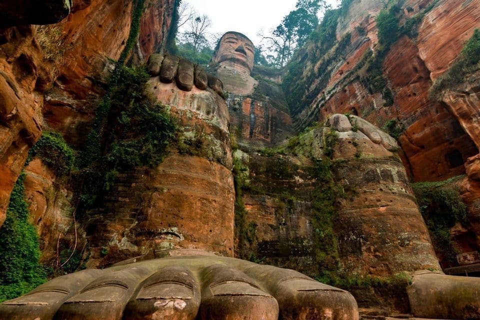 Una vista dal basso di una colossale statua di Buddha scolpita in una scogliera rossa, con i suoi grandi piedi in primo piano.