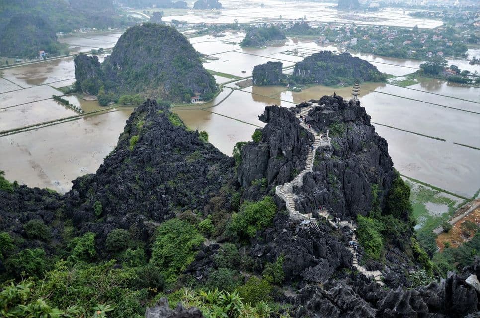 Una escalera de piedra serpentea por una escarpada montaña de roca oscura, con vistas a un vasto paisaje de arrozales inundados y otras formaciones kársticas.