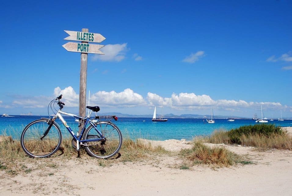 Ein Fahrrad steht neben einem hölzernen Wegweiser an einem Sandstrand mit Blick auf ein türkisfarbenes Meer mit Segelbooten.