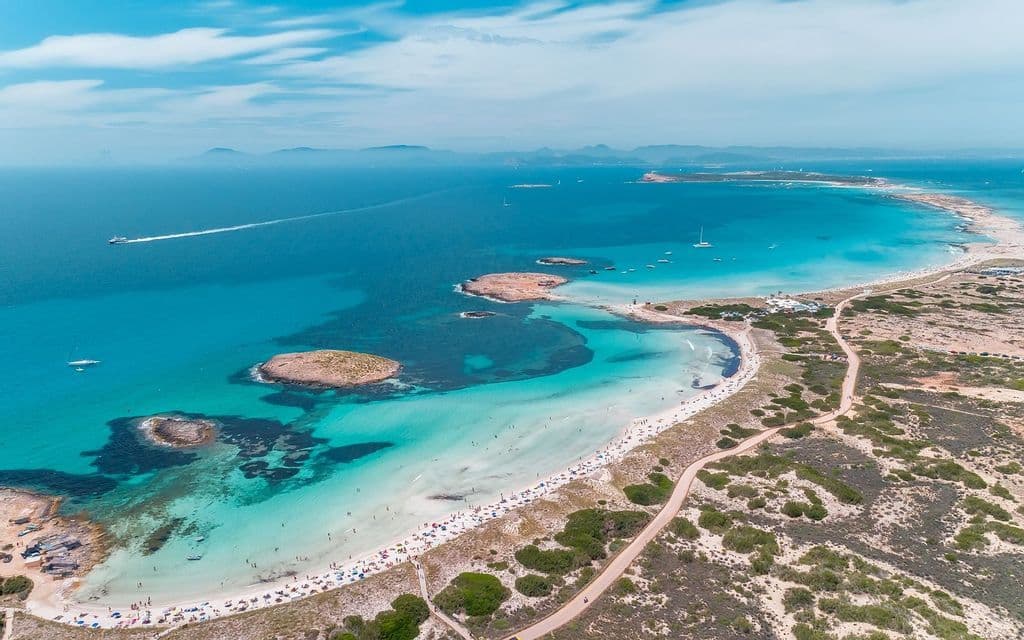 Una vista aerea di una lunga spiaggia di sabbia curva, affollata di gente, che lambisce un mare turchese con barche e piccole isole.
