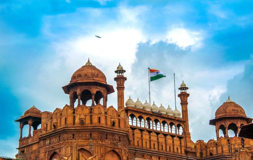 Die indische Flagge weht über einer verzierten roten Sandsteinfestung mit mehreren Kuppeln und Türmen vor einem bewölkten blauen Himmel.