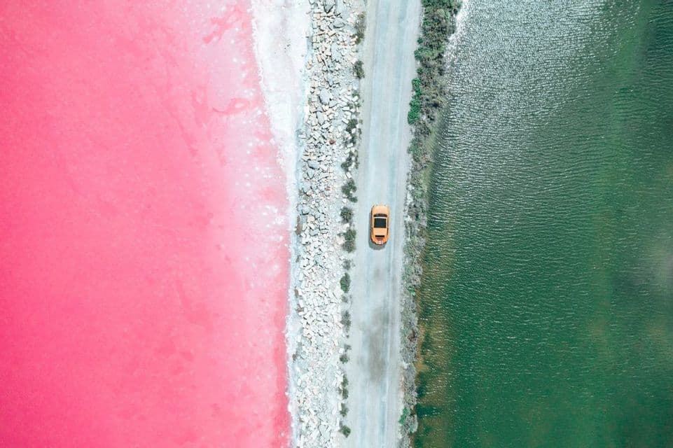Una vista aerea di un'auto gialla che guida su una strada che separa un lago rosa vibrante da uno verde scuro.
