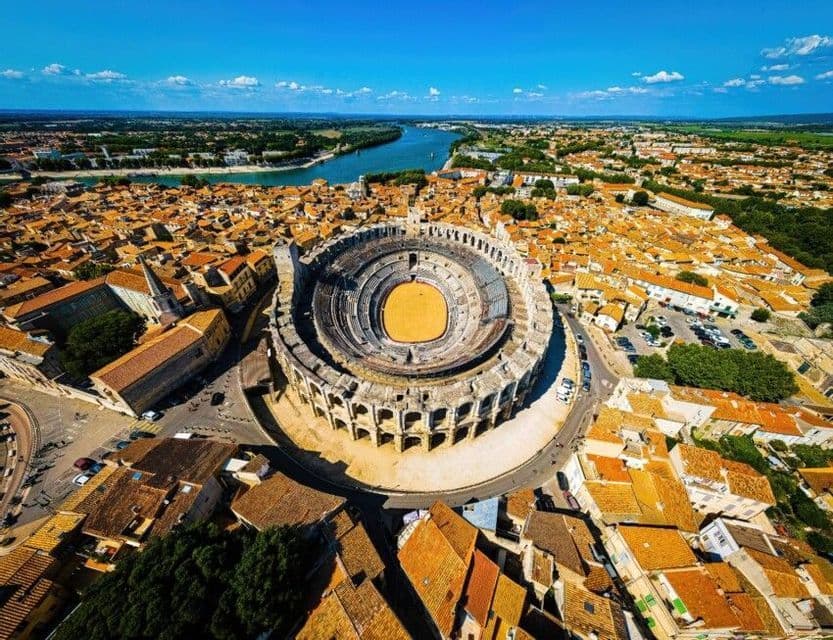 Veduta aerea di un grande anfiteatro in pietra al centro di una città con tetti in terracotta, con un fiume in lontananza sotto un cielo azzurro.