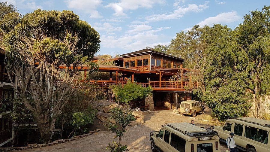 Safari jeeps are parked outside a multi-level wooden lodge surrounded by lush trees and rocks under a partly cloudy sky.