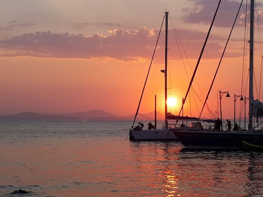 Diverse barche a vela ormeggiate in un porto, sagomate contro un tramonto arancione che si riflette sull'acqua.