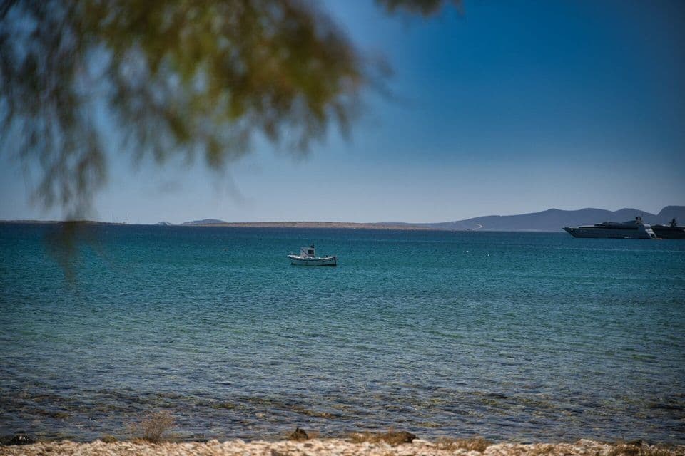 Una piccola barca bianca galleggia su acque tranquille e turchesi, con un'isola lontana e uno yacht sullo sfondo sotto un cielo azzurro.
