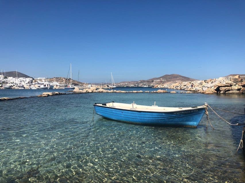 Una piccola barca blu galleggia in acque limpide e turchesi vicino a una costa rocciosa, con un porto e una città in lontananza sotto un cielo azzurro.