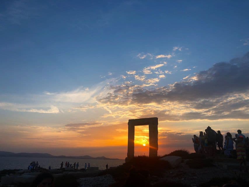Un viaggio di gruppo WeRoad osserva il sole tramontare attraverso un grande arco di pietra in riva al mare sotto un cielo colorato.