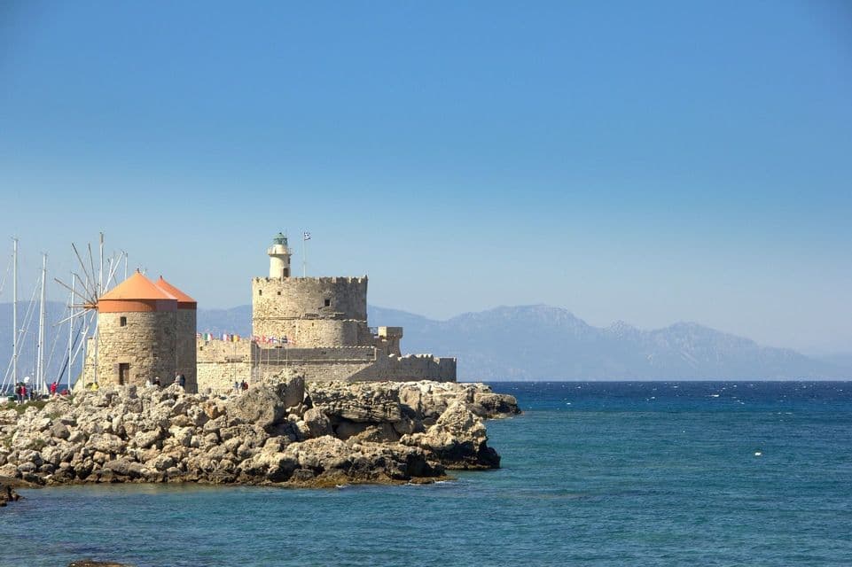 Un mulino a vento in pietra con tetto arancione e un faro fortificato si ergono su una costa rocciosa accanto al mare blu, con montagne in lontananza.