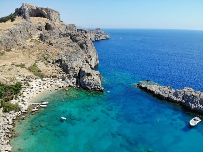 Una vista aerea di una piccola spiaggia incastonata in una cala rocciosa, con rovine sulla scogliera e barche nel mare limpido e turchese.