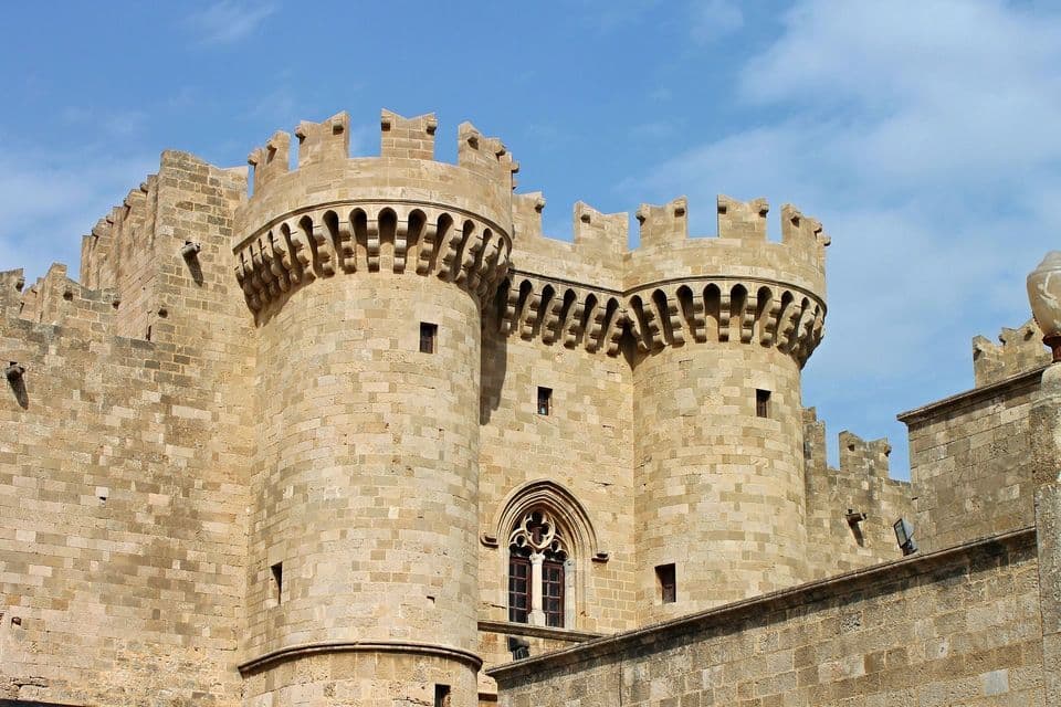 Due grandi torri rotonde di un castello in pietra con merlature, sotto un cielo azzurro con nuvole leggere.