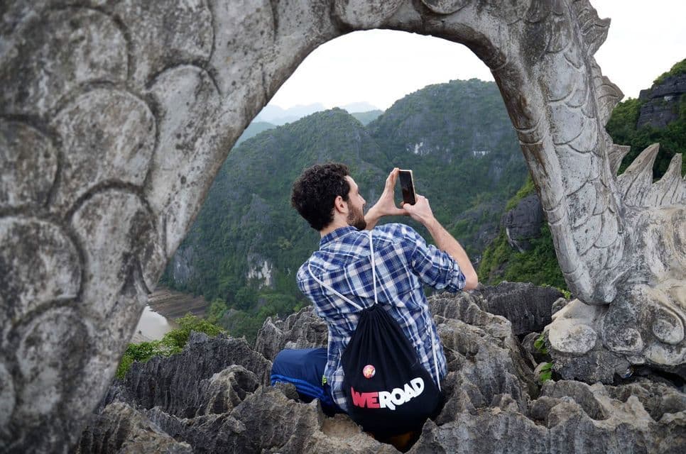 A man with a WeRoad backpack takes a photo with his phone of a mountain view, framed by a stone dragon sculpture.