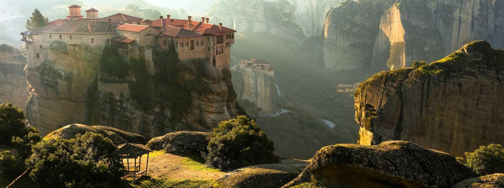 A stone monastery with a red-tiled roof perched on a sheer rock cliff, with another visible in the hazy background valley.