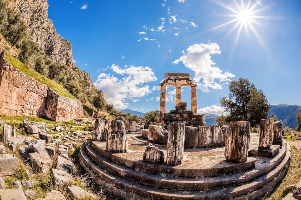 Las ruinas de un antiguo templo circular con columnas de piedra en la ladera de una montaña bajo un sol brillante y cielo azul.