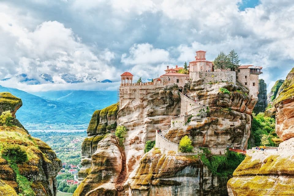 A stone monastery built on a sheer rock pillar, with a winding staircase and a view over a vast mountain valley under a cloudy sky.